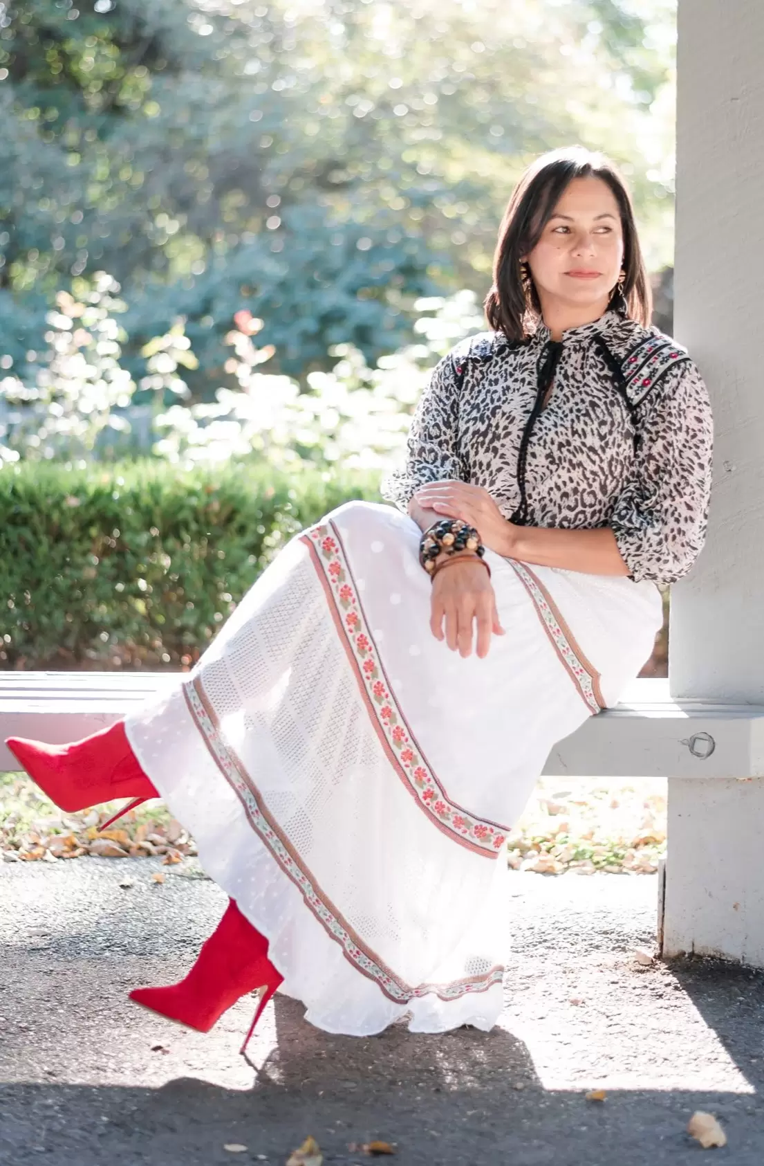 A woman with a positive mindset sits on a bench outdoors, wearing a leopard print top, long white skirt with red accents, and red boots.